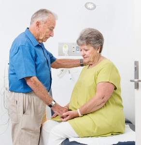 An older man holding the hat of an older lady seated on a hospital bed.