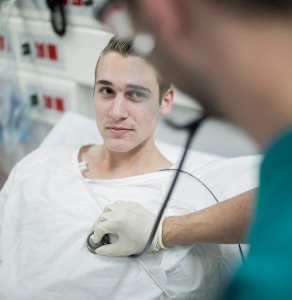 A young man lying in a gown in a hospital bed as a health professional not in focus places a device on his heart.