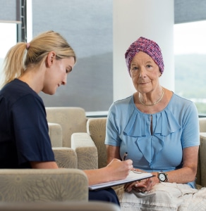 A health professional with a pen and clipboard in hand filing out a form as they are sat with an older cancer patient.