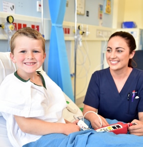 A young boy smiling while lying on a hospital bed as a nurse smiles at him.