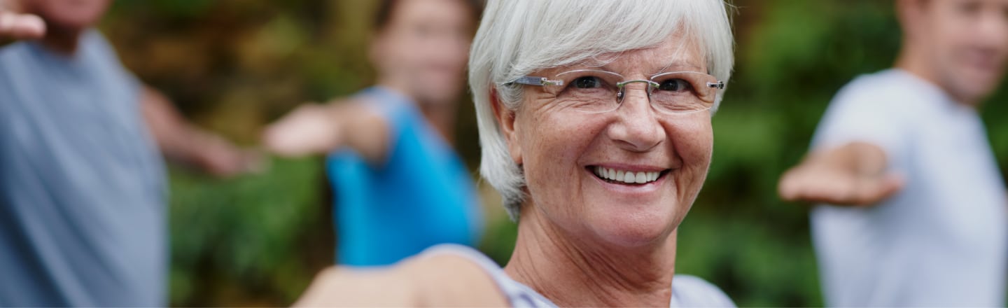 An older adult smiling while outside doing balance exercises with other adults in the background out of focus.