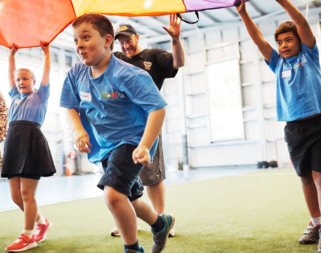 A young boy running under a parachute while an adult and two other children hold it up at a Go4Fun program session.