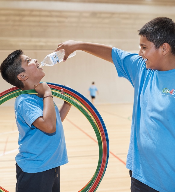 A boy sweating pouring water from a water bottle into another boy who is holding hoola hoops mouth at a Go4Fun session.. Both are wearing Go4Fun t-shirts.