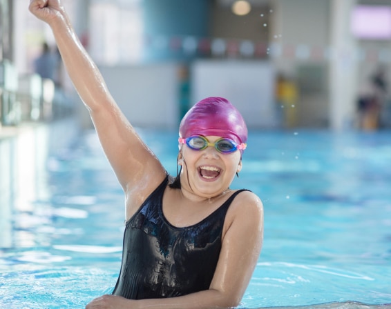 A girl in an indoor swimming pool raises her one arm aloft in celebration as she rests on the side of the pool.