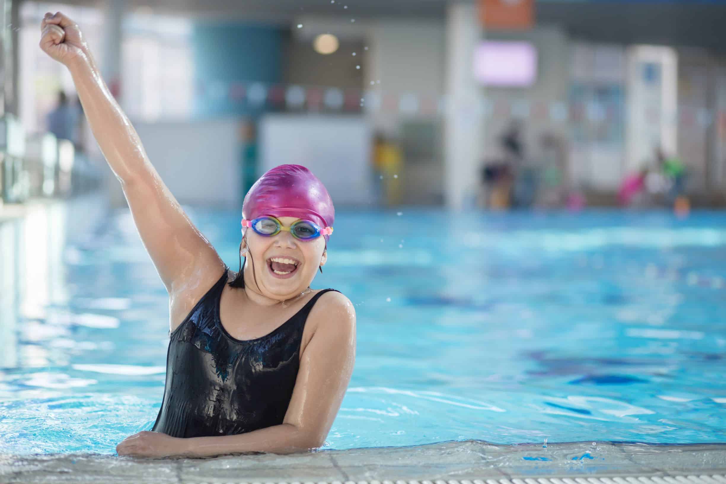 A girl in an indoor swimming pool raises her one arm aloft in celebration as she rests on the side of the pool.