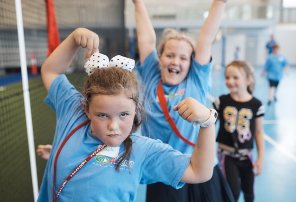 A young girl wearing a Go4Fun t-shirt flexing her muscles at the camera while another girl celebrates in the background at a Go4Fun session.