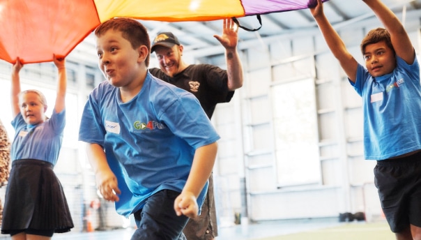 A young boy running under a parachute while an adult and two other children hold it up at a Go4Fun program session.