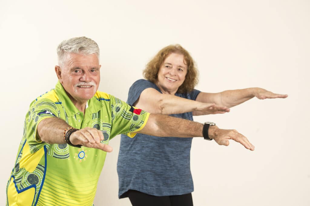 Two Aboriginal older adults doing balance exercises in front of a plain background.
