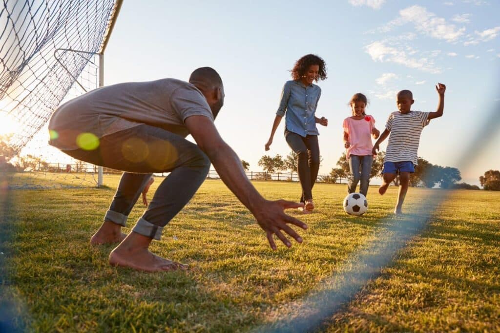 A mother, daughter and boy playing soccer about to kick a ball at goal as the father plays goalkeeper.