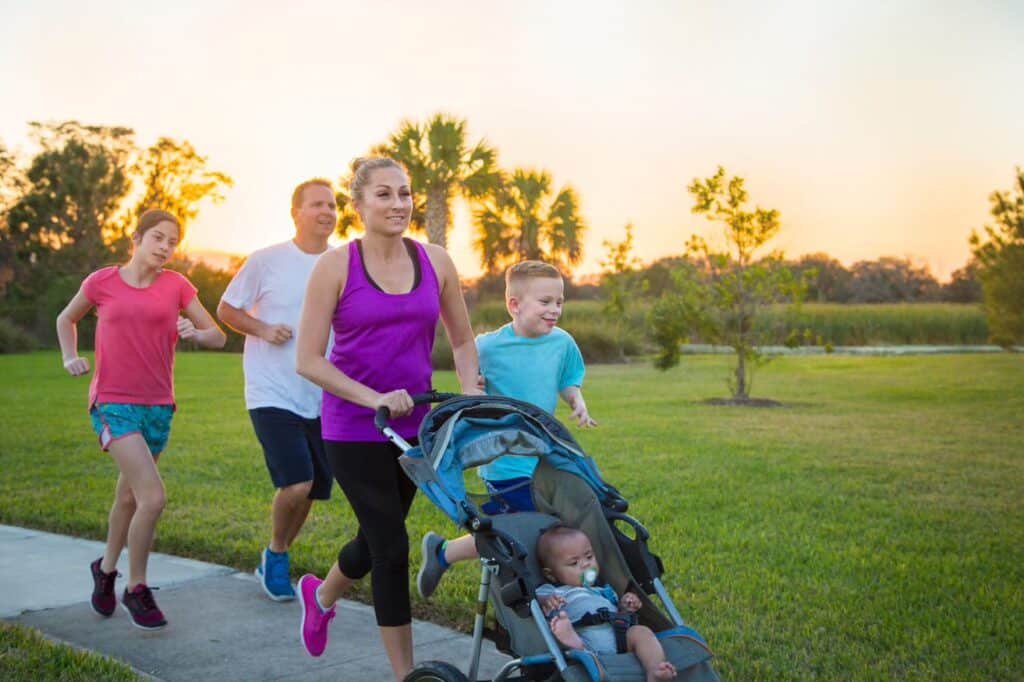 A family of five on a pathway. The mother, father, young son and young daughter are running, while the mother pushes a pram with a young child in.