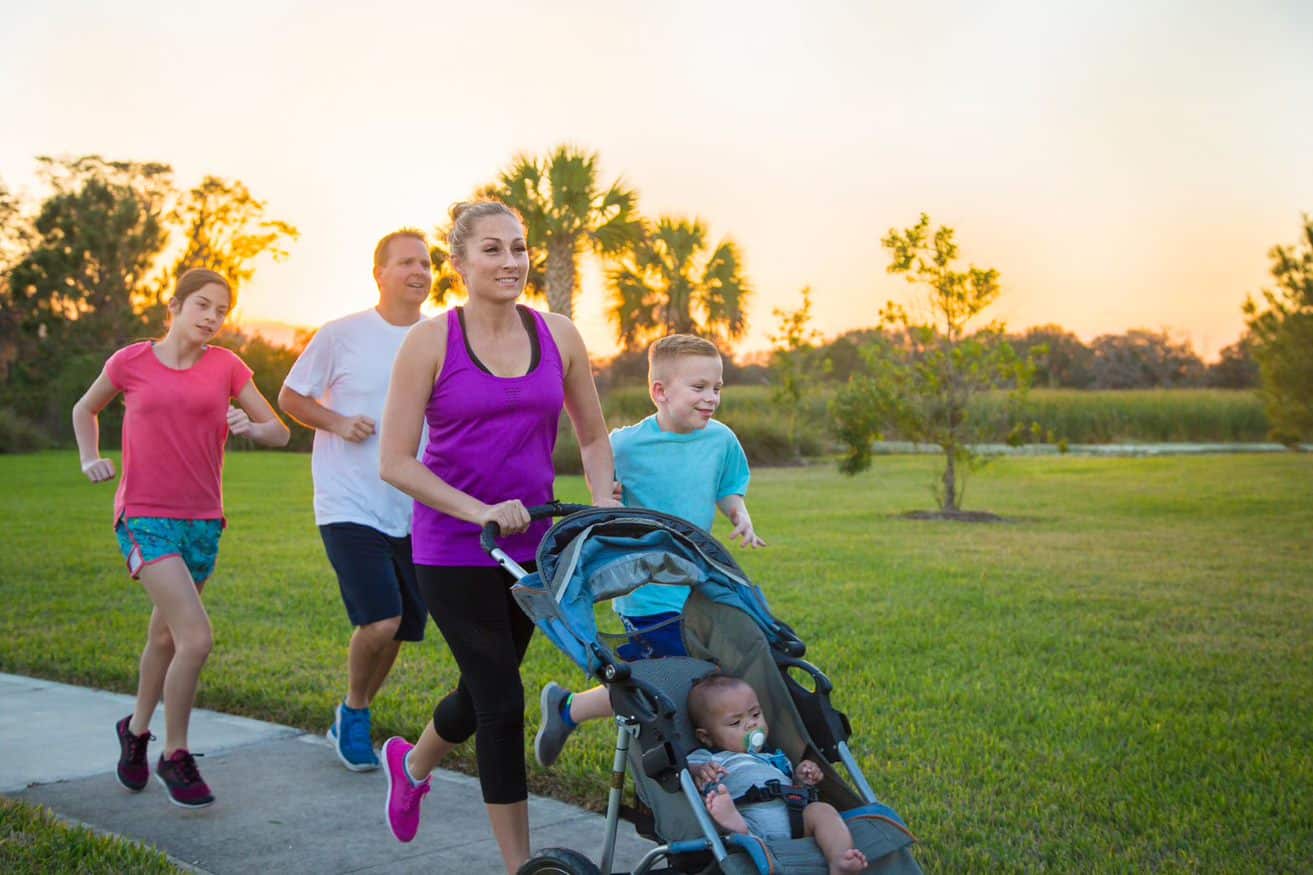 A family of five on a pathway. The mother, father, young son and young daughter are running, while the mother pushes a pram with a young child in.