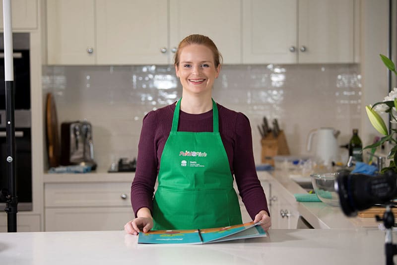 A young woman with an apron on smiling at a kitchen bench with the Patch to Plate recipe book open.