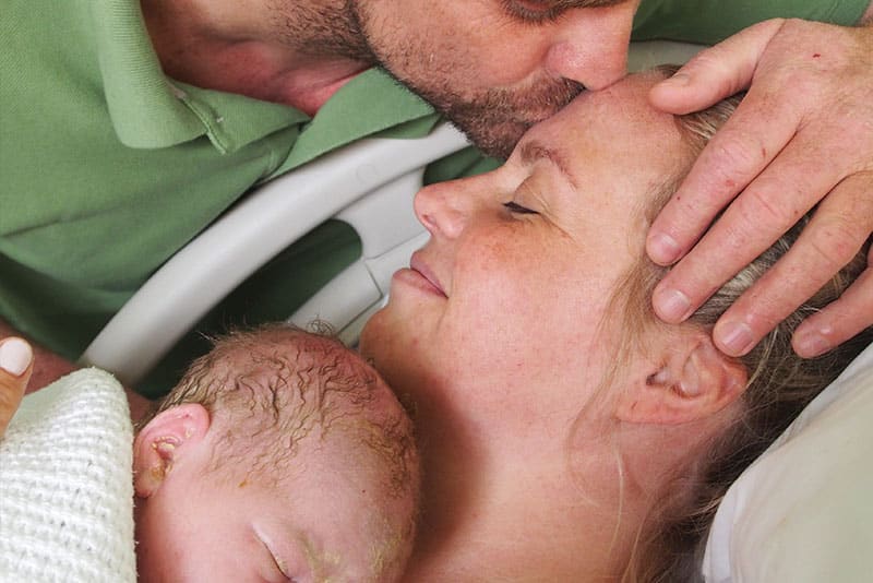 A mother holding her newborn as a father kisses her forehead.