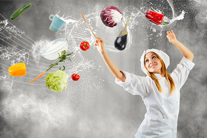 A smiling woman in a chef's outfit with healthy ingredients up in the air around her.