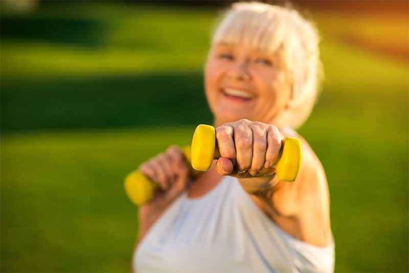 An older woman smiling while she holds a small dumbbell weight to the camera.