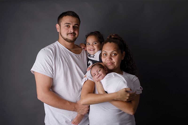 A mother holding her child with the father and young daughter also in shot behind a plain backdrop.