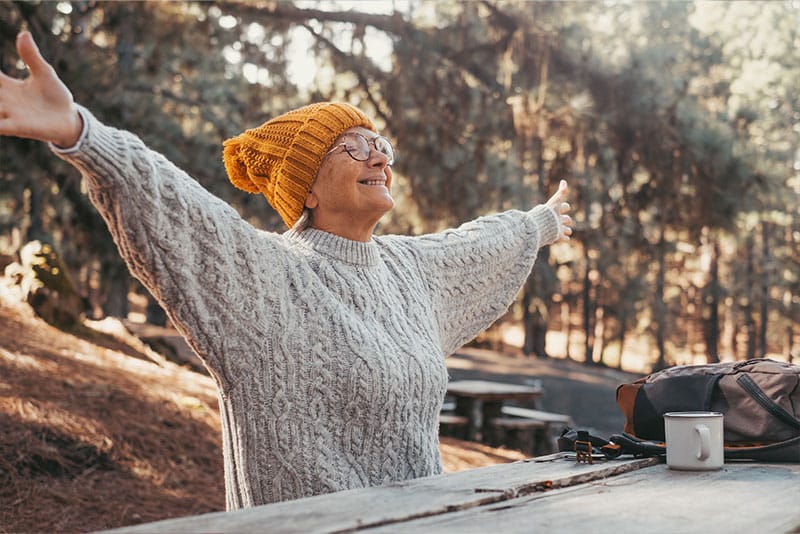 An older lady smiling with her eyes closed and arms stretched out taking in nature's surroundings while sat at a table in a woodland area.