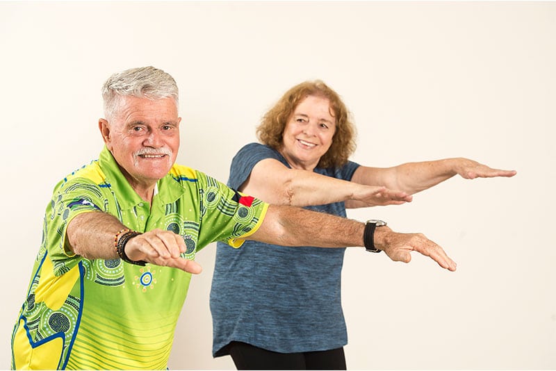 Two Aboriginal older adults doing balance exercises in front of a plain background.