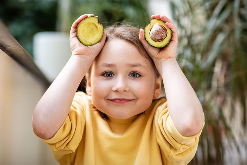 A young child holding an avocado above her head smiling.