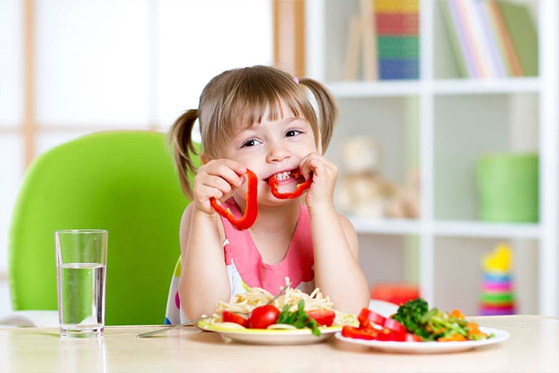 A smiling young girl eating capsicum from a plate of fruit and vegetables.