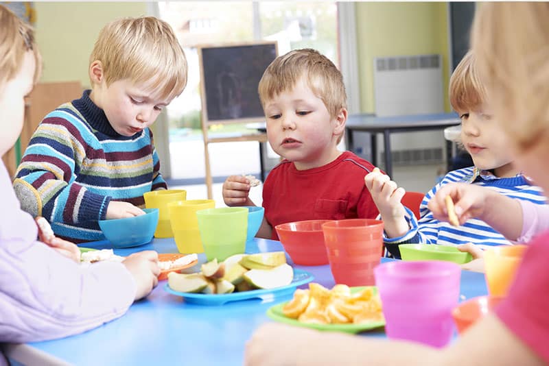 Young children at a table in an early childcare service with plastic cups and plates of fruit.