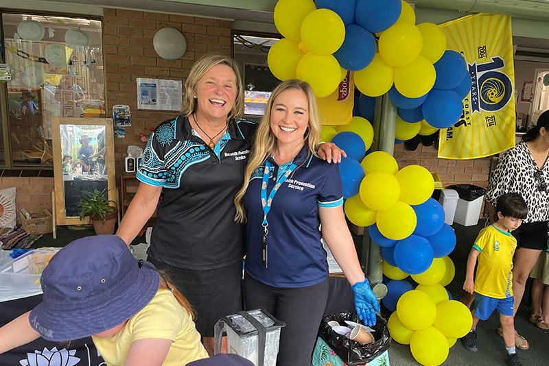 Two members of the Central Coast Health Promotion team smiling arm in arm while at an event with balloons in the background.