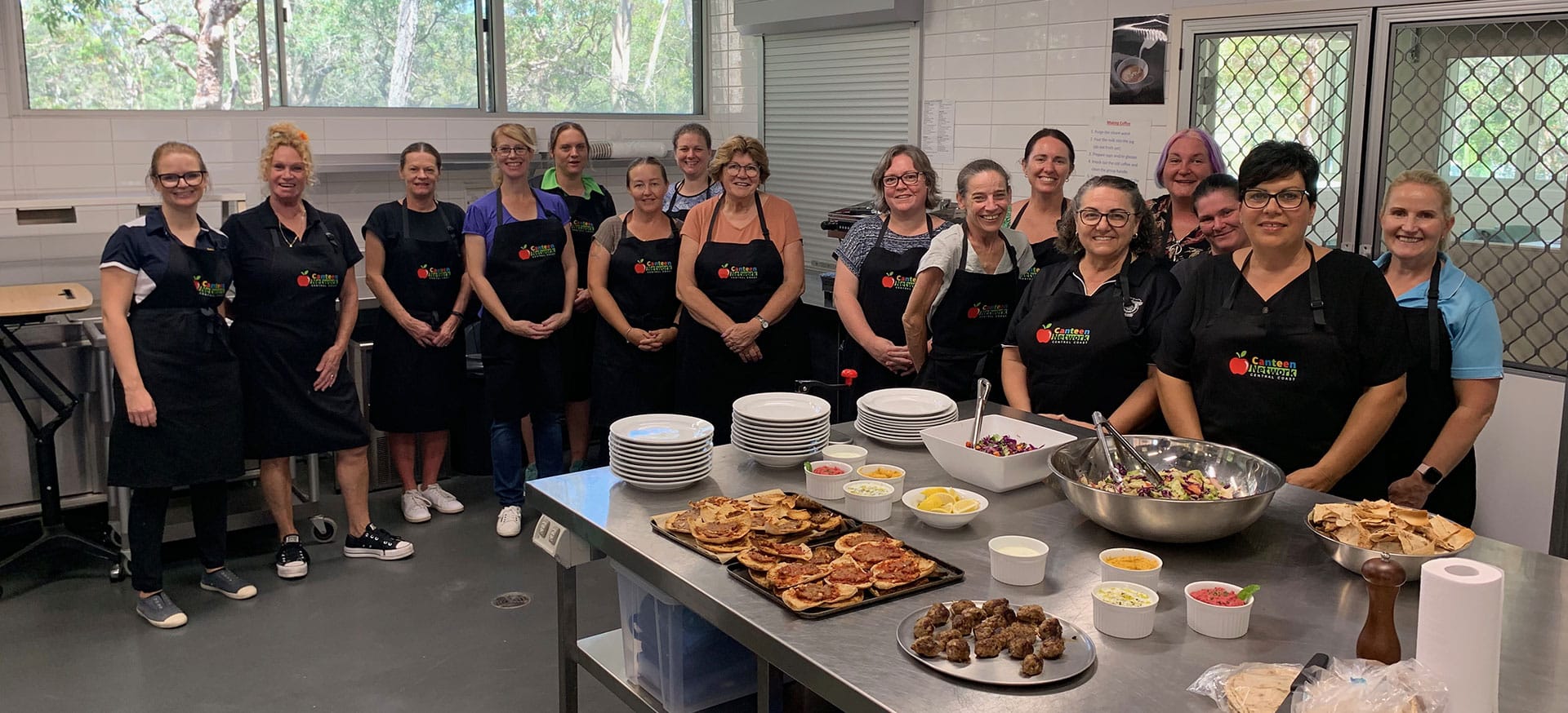 A group shot of canteen staff and Health Promotion and Nutrition Services staff at a Canteen Network meeting in a school kitchen with food on the table.