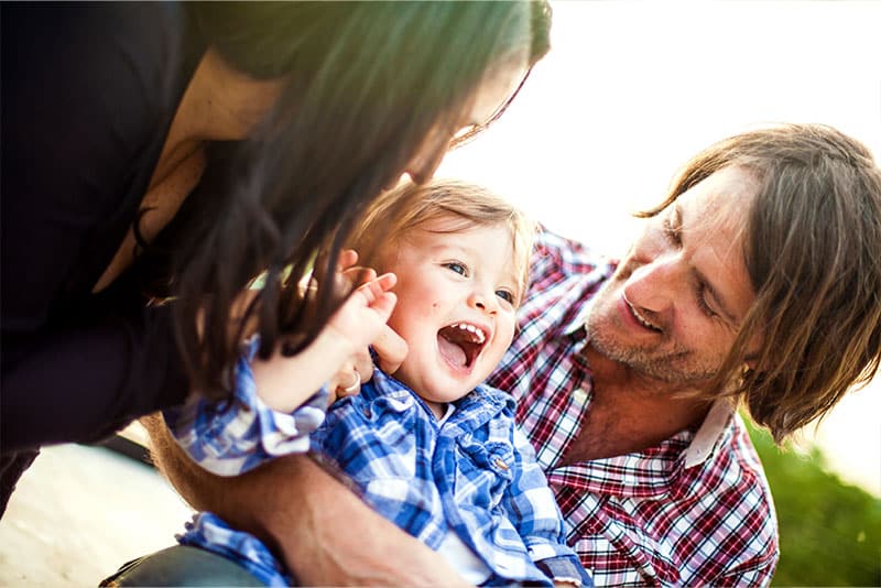 A mum and dad smiling while holding their young child outdoors.