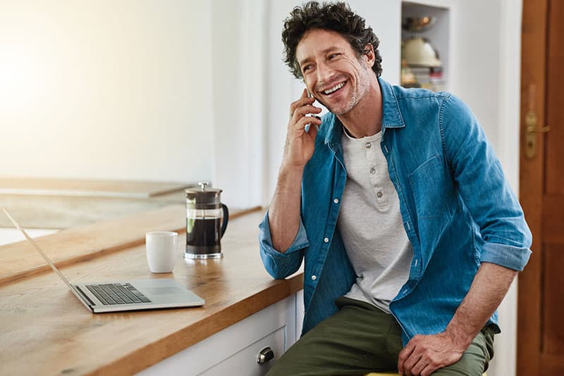 An adult man smiling while talking on his mobile phone at a table with his laptop open.