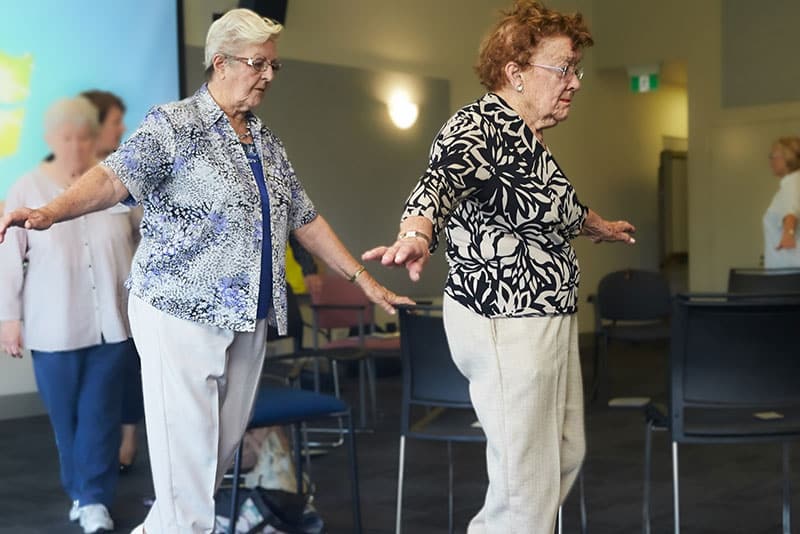 A group of older adults doing balance exercises while walking around a circle of chairs inside.