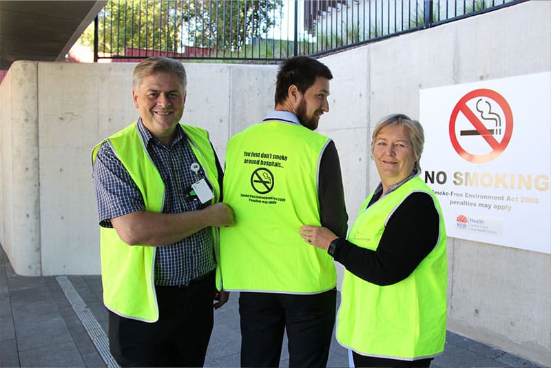 Three Health Promotion staff with high-vis jackets on, two of which are pointing at the back of the third person's jacket that says "You just don't smoke around hospitals" with a no smoking sign on it and another no smoking sign in the background.