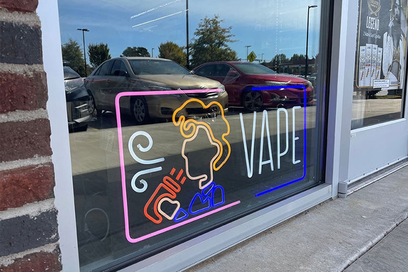 A vape shop with a neon sign in the window with the word VAPE and a silhouette of someone vaping.
