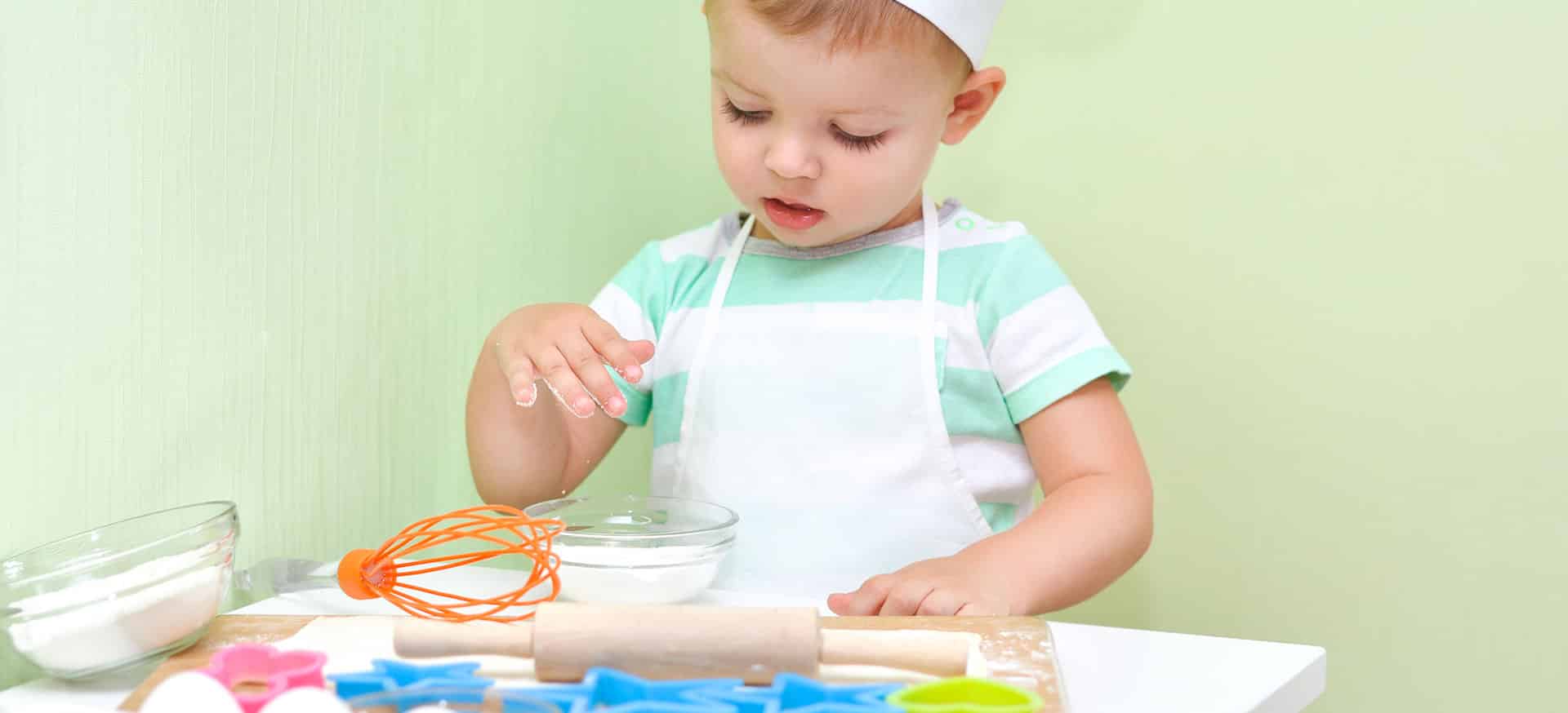 A child in an apron and hat with flour and a roller and rolling board.