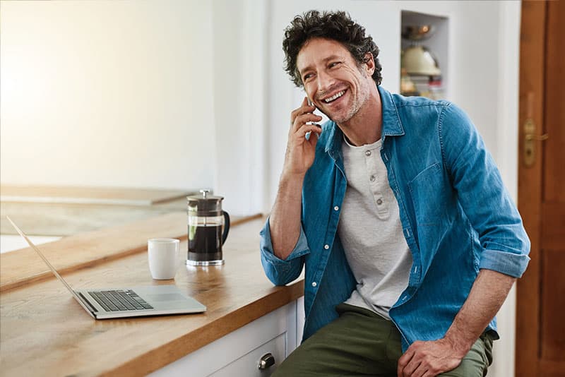 An adult man smiling while talking on his mobile phone at a table with his laptop open.