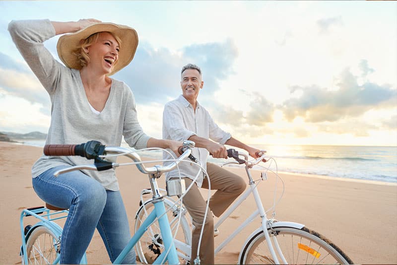 An adult man and adult woman smiling while cycling on a beach.