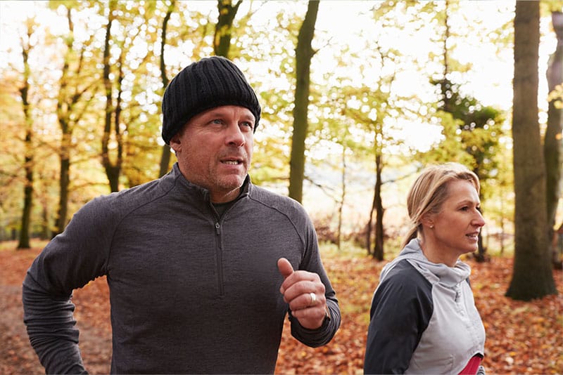 An adult man and woman running in a leafly woodland.