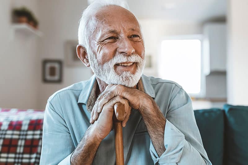 An older man smiling as he rests his hands and chin on his walking stick while on a sofa inside.