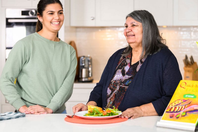 An Aboriginal grandaughter and grandmother at a kitchen bench with a plate of food in front of them and the Quick Meals for Kooris At Home manual on display.