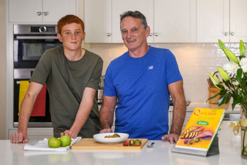An Aboriginal father and son at a kitchen bench with a plate of food in front of them and the Quick Meals for Kooris At Home manual on display.