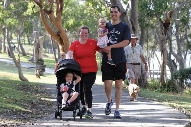 A family walking along a pathway with the mother pushing a young child in a pram and the dad holding another child. A man walks a dog in the background.