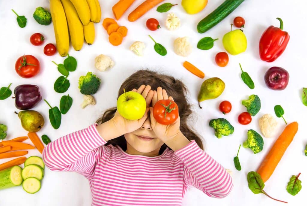 A young girl covers covers her face with fruit while other fruit and vegetables surround her.
