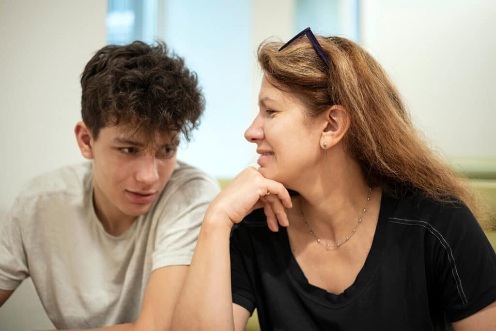 A middle-aged woman is spending time with her son in a cafe, they sit and talk. Mum looks at her teenage son with love.