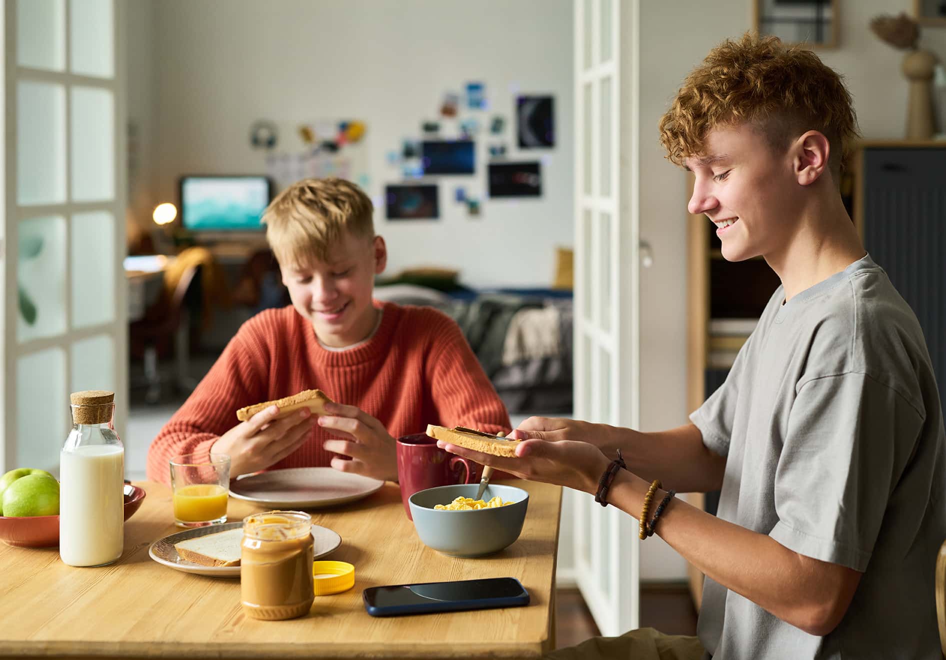 Two Caucasian teenage boys sitting at table eating breakfast together, smiling and holding toast, smartphone and breakfast foods visible on table, casual home setting