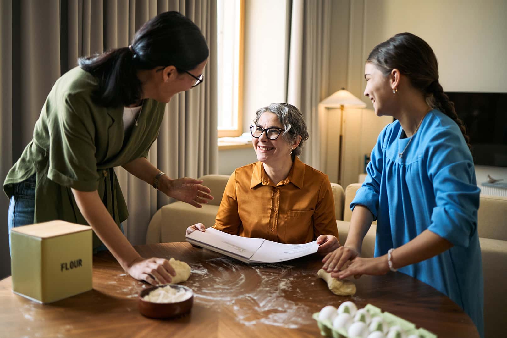 Senior woman reviewing recipe while middle aged woman and teenager kneading dough at table. Family bonding and home cooking concept for caregiving, lifestyle, support marketing
