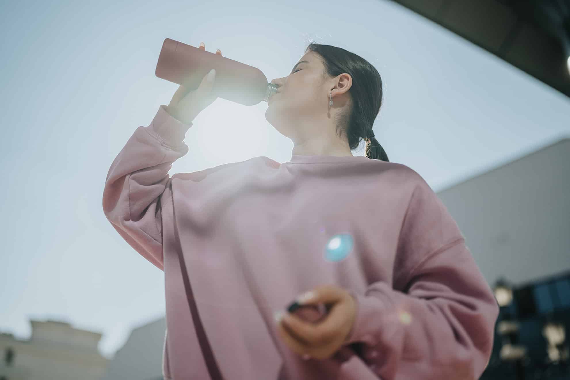 Young woman drinks from a reusable water bottle while standing outdoors. A low-angle view shows her in a pink sweatshirt hydrating in sunlight.