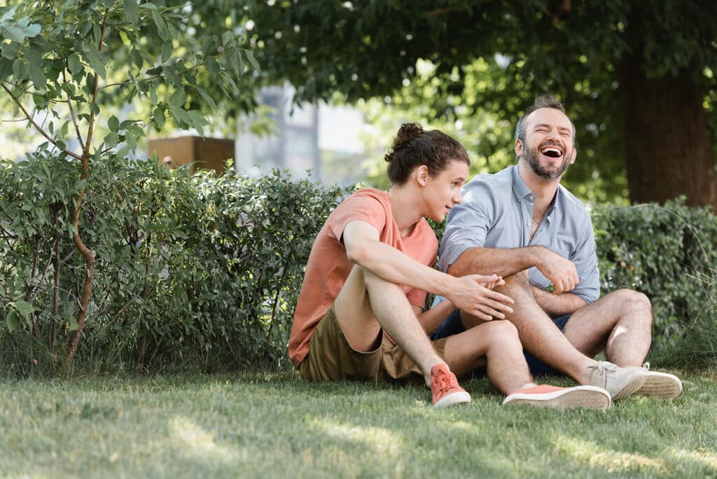happy father laughing while sitting near teenage son on green lawn