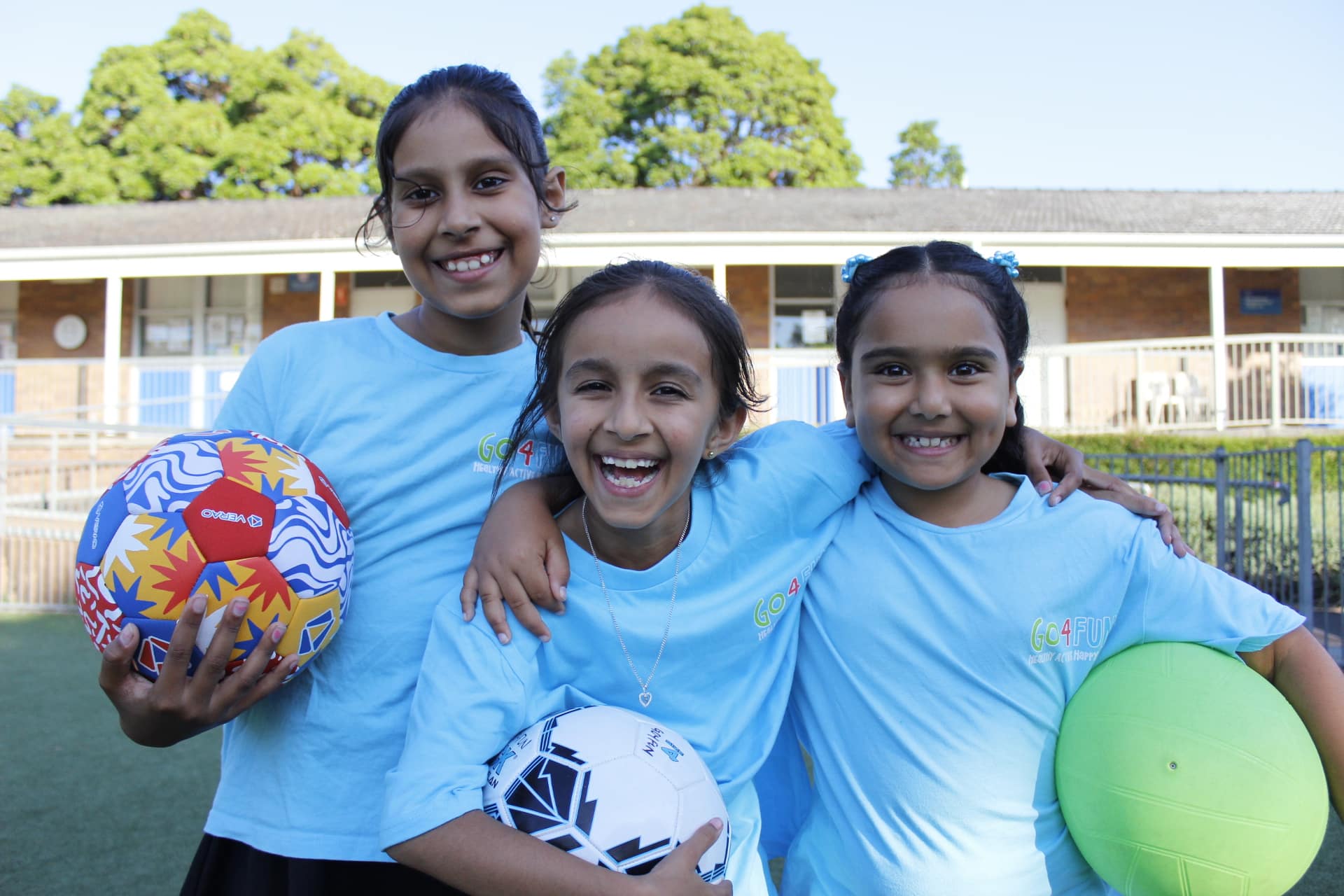 Group photo taken outside of three primary school-aged girls smiling, looking at the camera and holding soccer balls.